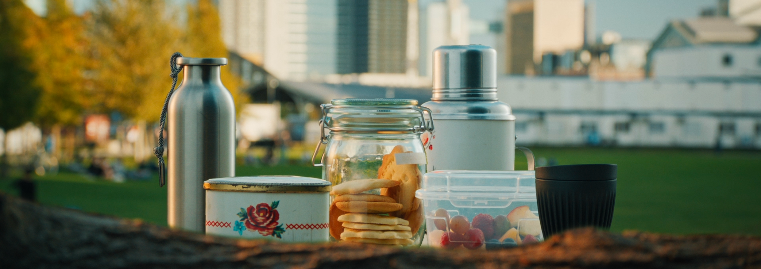 Een roestvrijstalen drinkfles, een roestvrijstalen thermos, een glazen bokaal, een blikken koekjestrommel, een plastic bewaardoosje en een herbruikbare beker op een tafel in een park.