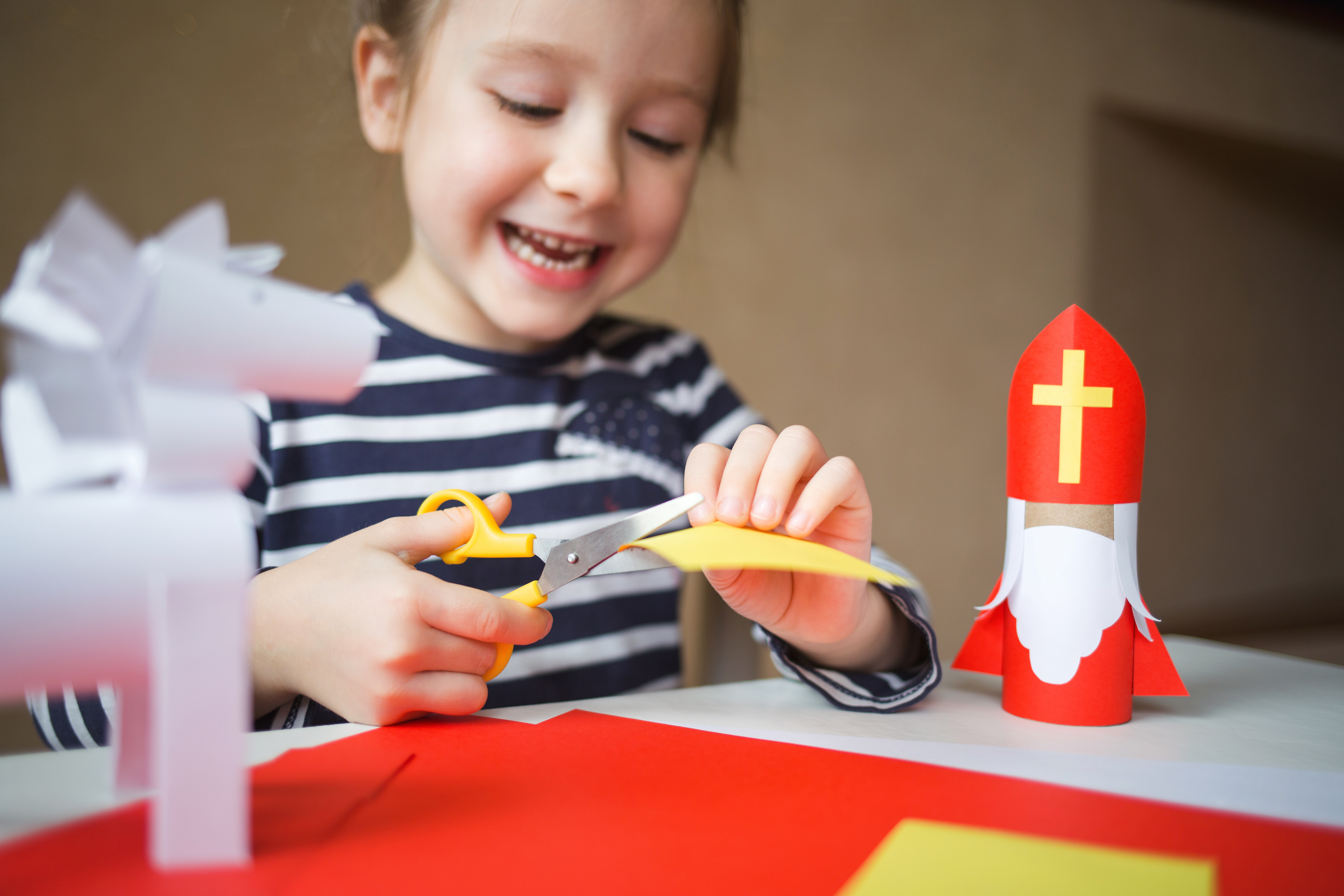 Een meisje knutselt Sinterklaas uit een leeg toiletrolletje en gekleurd papier. Ze knipt met een schaar in een stukje geel papier.